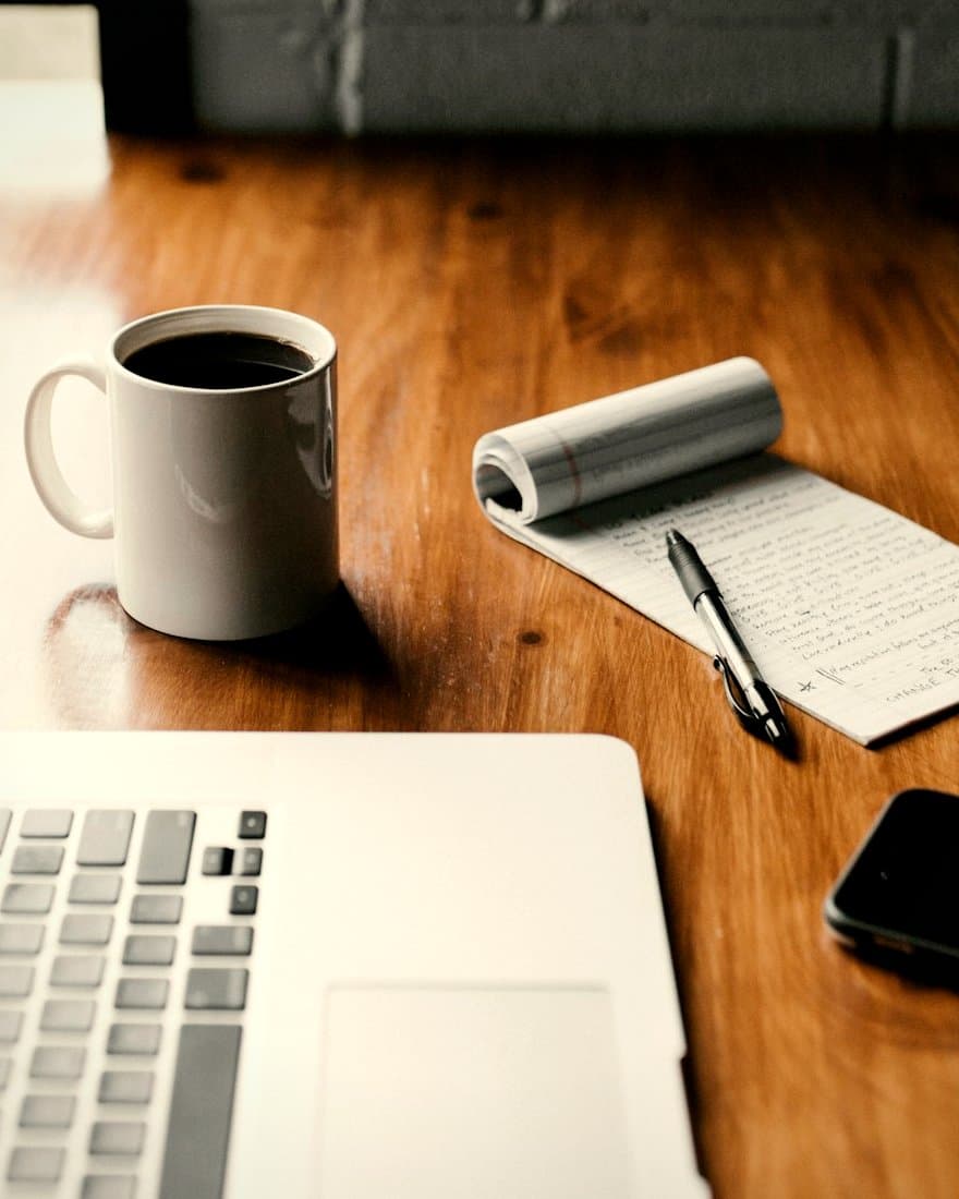 Mesa de madera cálida con una taza de café, una libreta con notas escritas a mano y un bolígrafo, un portátil y un móvil. Imagen de ejemplo.
