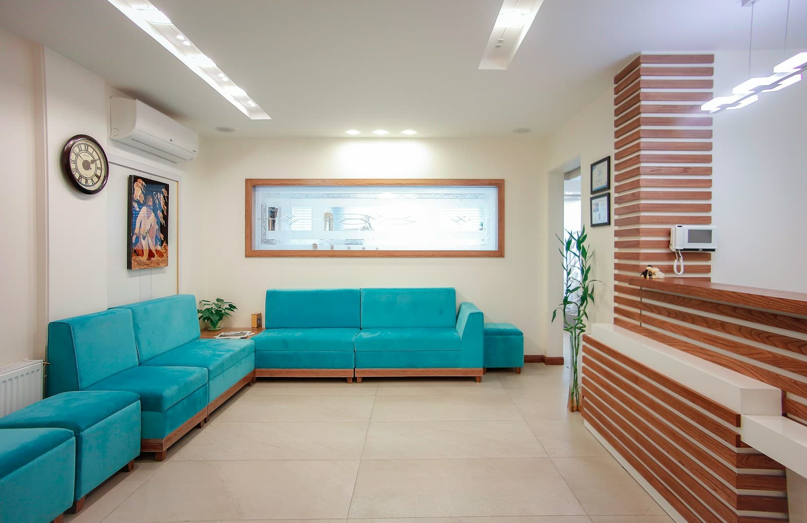 Sala de espera luminosa de una clinica de fisioterapia, con sofas en tono verde azulado, pared de madera clara en la zona de recepcion y una planta. Imagen de muestra.
